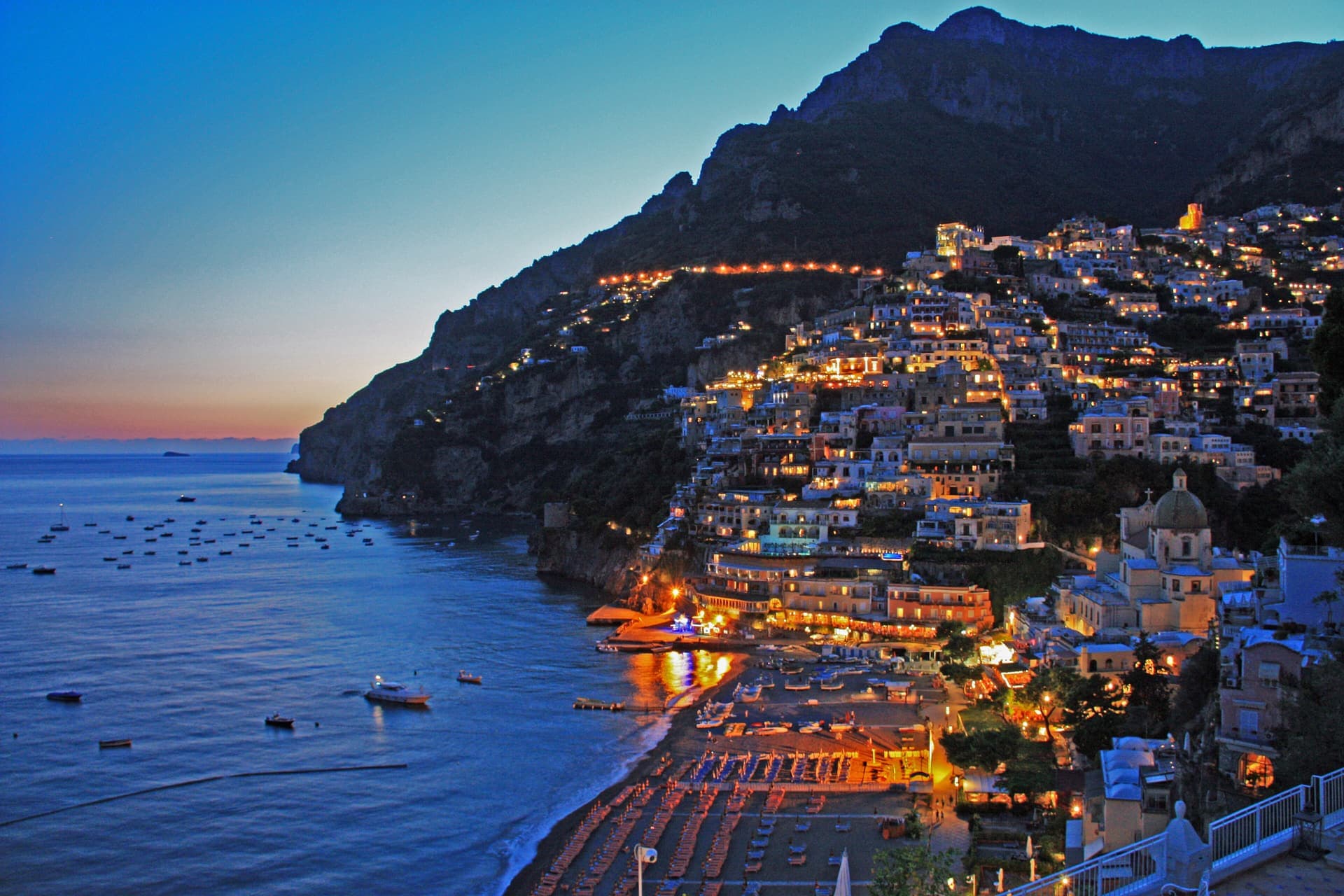 Positano at dusk, with lit houses climbing the hillside above a blue bay